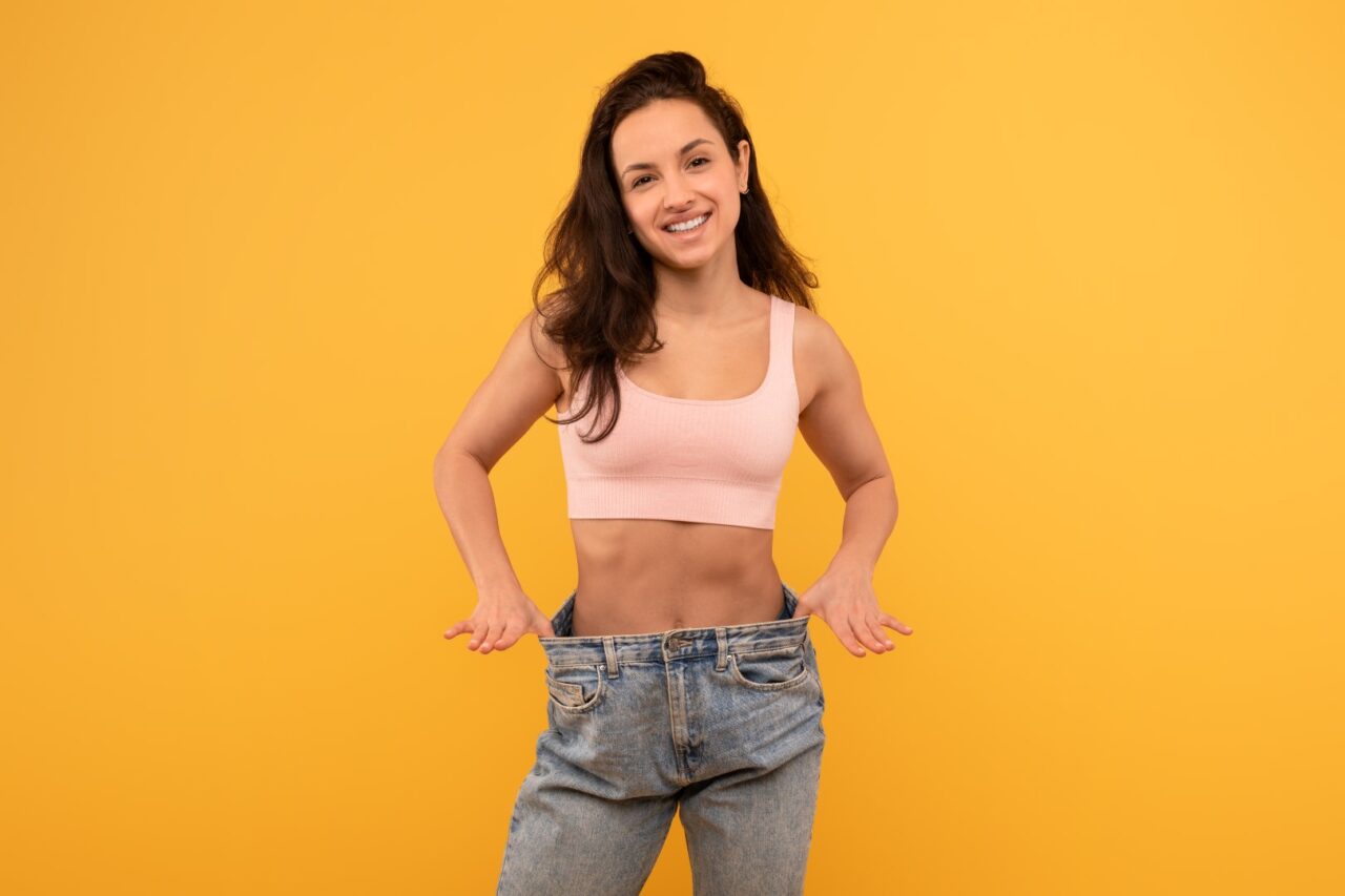 A cheerful young woman stands with hands on her hips wearing a light pink crop top and oversized blue denim jeans. She smiles confidently against a bright yellow backdrop